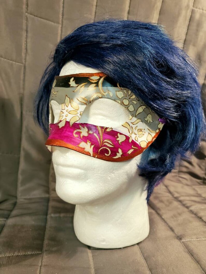 K&ouml;nnte beinhalten: Eine dekorative Augenmaske mit einem floralen Muster in Wei&szlig;, Gold und Pink. Die Maske befindet sich auf einem wei&szlig;en Schaufensterkopf mit blauen Haaren. Die Maske hat ein mehrschichtiges Design mit Metallic-Finish.