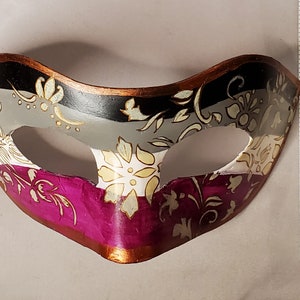 K&ouml;nnte beinhalten: Eine dekorative Maske mit floralem Design. Die Maske hat ein Farbschema aus Schwarz, Grau, Wei&szlig; und Magenta mit goldenen Akzenten. Die Maske ist etwa 18 cm breit.