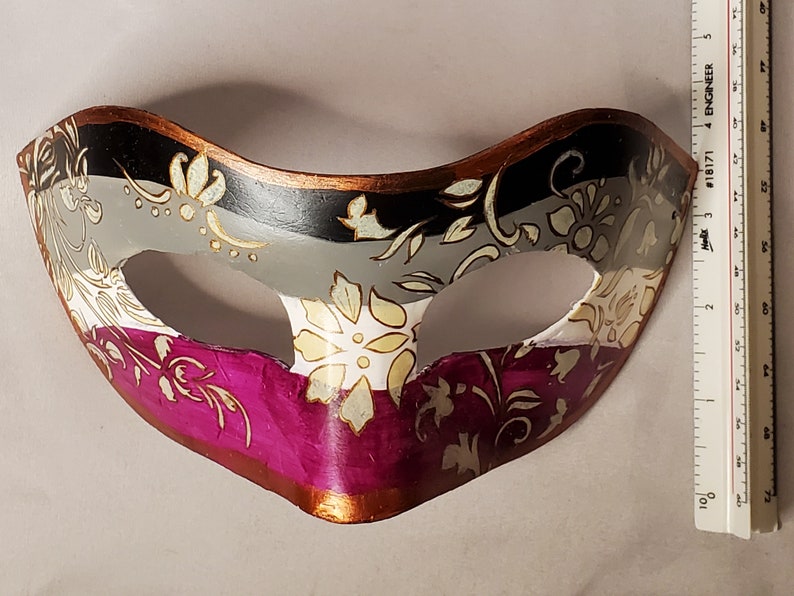K&ouml;nnte beinhalten: Eine dekorative Maske mit floralem Design. Die Maske hat ein Farbschema aus Schwarz, Grau, Wei&szlig; und Magenta mit goldenen Akzenten. Die Maske ist etwa 18 cm breit.