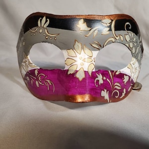 K&ouml;nnte beinhalten: Eine dekorative Maskenballmaske mit einem Farbblock-Design. Der obere Bereich ist schwarz, grau und wei&szlig;, w&auml;hrend der untere Bereich ein leuchtendes Magenta ist. Goldene Blumenmuster zieren die Maske. Die R&auml;nder sind goldfarben.
