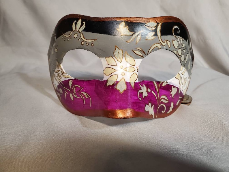 K&ouml;nnte beinhalten: Eine dekorative Maskenballmaske mit einem Farbblock-Design. Der obere Bereich ist schwarz, grau und wei&szlig;, w&auml;hrend der untere Bereich ein leuchtendes Magenta ist. Goldene Blumenmuster zieren die Maske. Die R&auml;nder sind goldfarben.