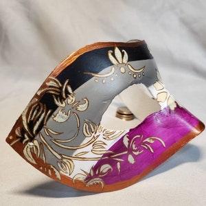 K&ouml;nnte beinhalten: Eine dekorative Maske in Schwarz, Grau, Wei&szlig; und Lila. Sie hat ein goldenes und braunes Blumenmuster. Die R&auml;nder sind in Braun und Gold eingefasst. Diese Maske ist wahrscheinlich f&uuml;r eine Maskerade oder Kost&uuml;mveranstaltung gedacht.