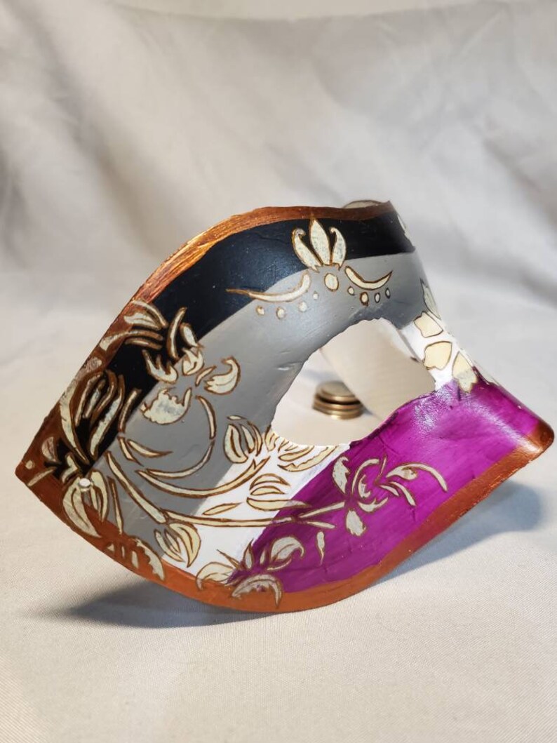 K&ouml;nnte beinhalten: Eine dekorative Maske in Schwarz, Grau, Wei&szlig; und Lila. Sie hat ein goldenes und braunes Blumenmuster. Die R&auml;nder sind in Braun und Gold eingefasst. Diese Maske ist wahrscheinlich f&uuml;r eine Maskerade oder Kost&uuml;mveranstaltung gedacht.
