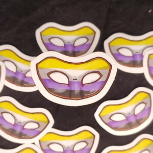 May include: Multiple stickers of a purple, yellow, and black genderqueer pride flag in the shape of a masquerade mask.