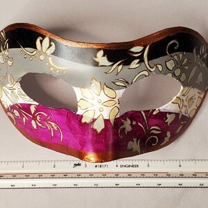 K&ouml;nnte beinhalten: Eine dekorative Maske mit floralem Design. Die Maske ist in drei Abschnitte unterteilt: Schwarz, Grau und Magenta. Goldene Blumenmuster zieren die Oberfl&auml;che. Die Maske hat einen braunen Rand und ist etwa 18 cm breit.