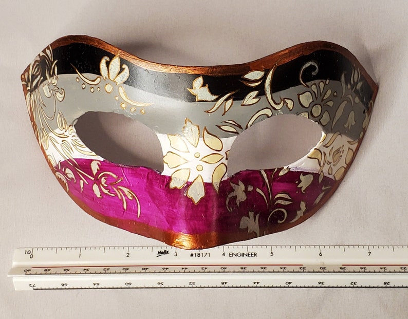 K&ouml;nnte beinhalten: Eine dekorative Maske mit floralem Design. Die Maske ist in drei Abschnitte unterteilt: Schwarz, Grau und Magenta. Goldene Blumenmuster zieren die Oberfl&auml;che. Die Maske hat einen braunen Rand und ist etwa 18 cm breit.