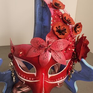 May include: A red and pink masquerade mask with a floral design. The mask is decorated with lilies, roses, and other flowers. The mask is on a blue stand.