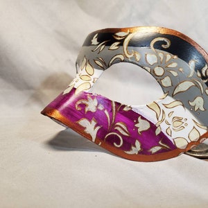 K&ouml;nnte beinhalten: Eine dekorative Maske mit einem bemalten Design. Die Maske hat ein Farbschema aus Schwarz, Grau, Wei&szlig; und Lila mit goldenen floralen Akzenten. Die R&auml;nder sind mit metallischer Goldfarbe verziert.