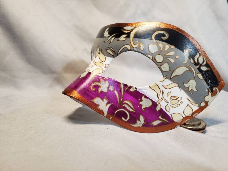 K&ouml;nnte beinhalten: Eine dekorative Maske mit einem bemalten Design. Die Maske hat ein Farbschema aus Schwarz, Grau, Wei&szlig; und Lila mit goldenen floralen Akzenten. Die R&auml;nder sind mit metallischer Goldfarbe verziert.