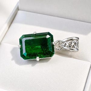 Puede incluir: Un colgante rectangular de piedra preciosa verde esmeralda con una fianza plateada. El colgante está engastado en un engaste de metal plateado y se muestra en una caja de joyería blanca.