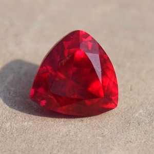 May include: A triangular, faceted, deep red gemstone. The polished surface of the gem has multiple facets that reflect light. The stone is a vibrant, saturated red colour, and appears to be a ruby. The gem is set against a neutral, textured background.