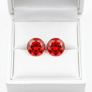 May include: Two round, faceted, bright red gemstones are displayed in a white jewelry box. The gems have a brilliant cut, reflecting light. The box is open, revealing the gems against a white interior.