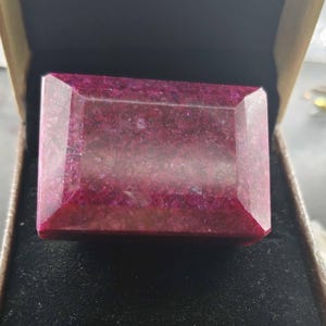 May include: A rectangular, faceted gemstone with a deep magenta and dark red hue, featuring a complex internal structure with visible inclusions and a polished surface, presented inside a small, open, tan-colored box with a black interior lining.