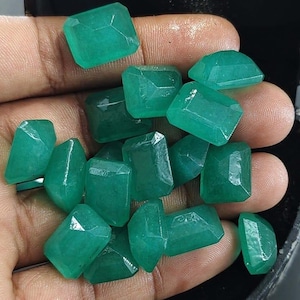May include: A collection of multiple loose, emerald-green gemstones with a rectangular, faceted cut. The stones exhibit a translucent quality and are displayed in a cluster, showing various angles of their polished surfaces and geometric shapes.