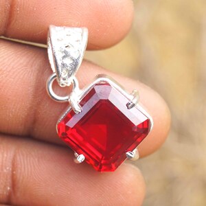 Puede incluir: Un colgante de plata con una llamativa piedra preciosa roja de talla cuadrada. La piedra preciosa está engastada en un marco de plata con una fianza texturizada. El colgante está sostenido por una mano.
