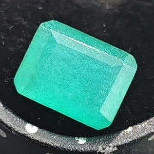 May include: A rectangular emerald-cut gemstone with a vibrant green hue and faceted surfaces that reflect light. The stone features sharp, clean edges and a polished finish, showcasing a consistent, saturated green color throughout its structure.