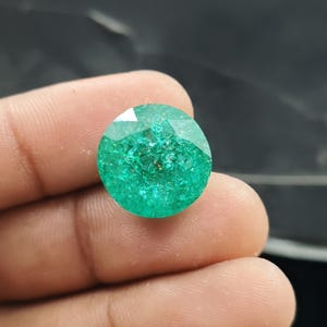 May include: A round, faceted gemstone with a vibrant emerald green colour and a highly textured, cracked internal appearance. The stone features a brilliant cut with multiple triangular facets that reflect light across its surface.