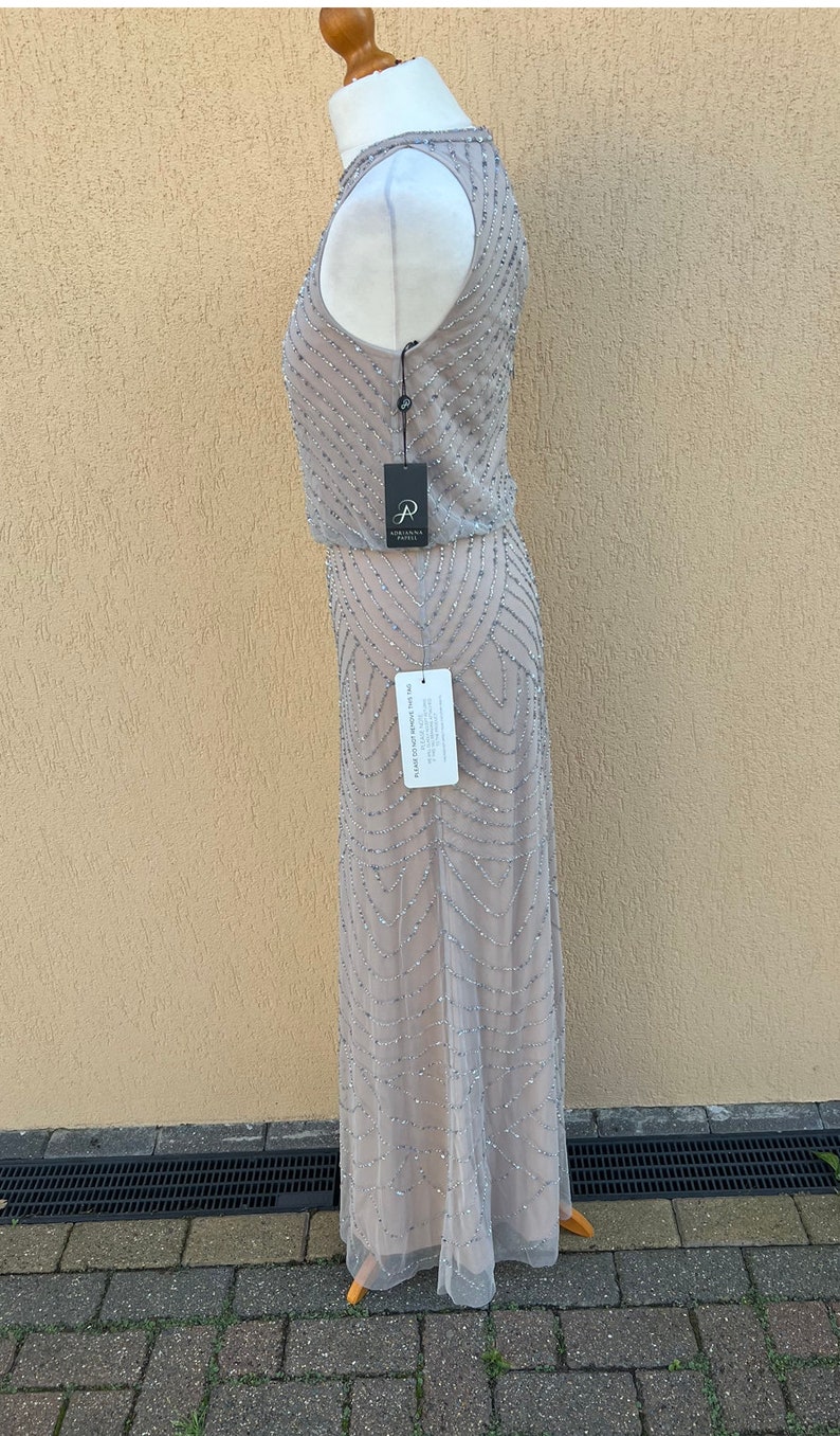 May include: A silver sequined maxi dress with a sleeveless design. The dress has a fitted bodice and a flowing skirt. The dress is on a mannequin and is displayed against a light beige wall.