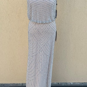 May include: A sleeveless, floor-length, beaded gown in a light gray color. The gown has a halter neckline and a geometric pattern.