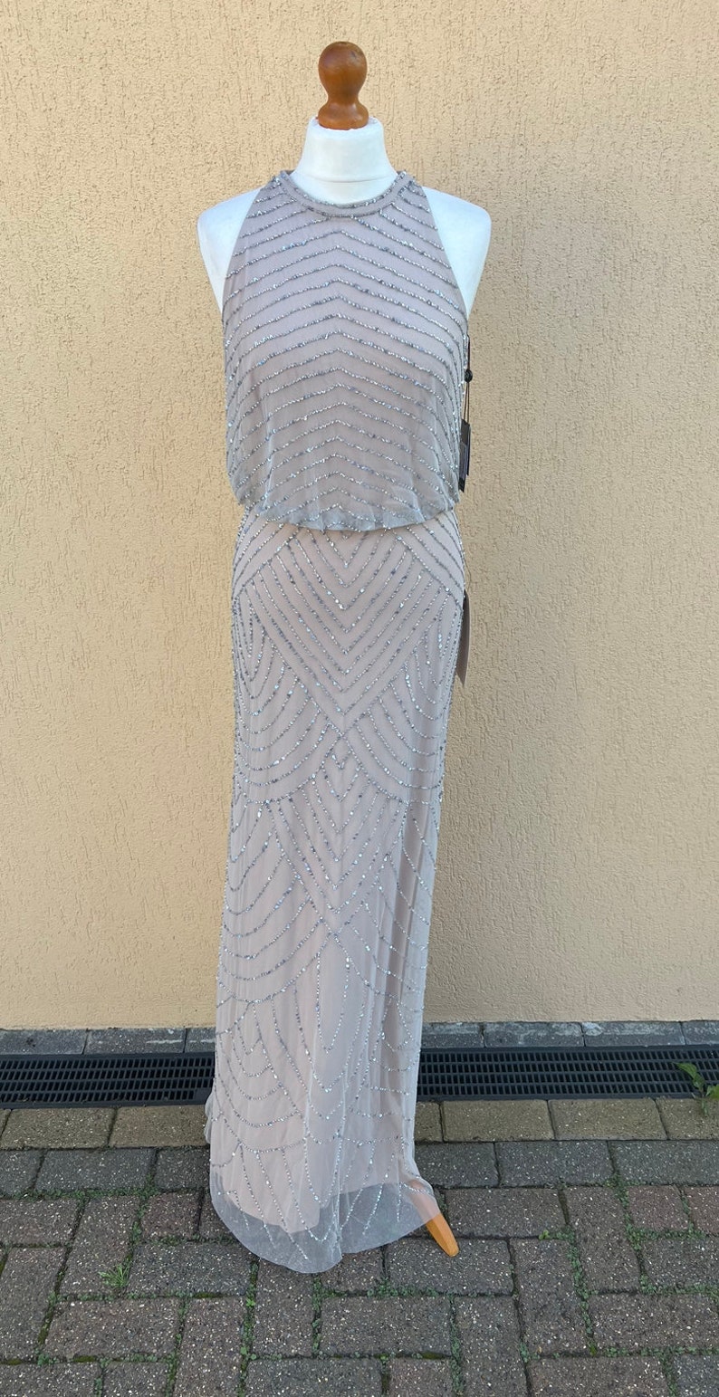 May include: A sleeveless, floor-length, beaded gown in a light gray color. The gown has a halter neckline and a geometric pattern.