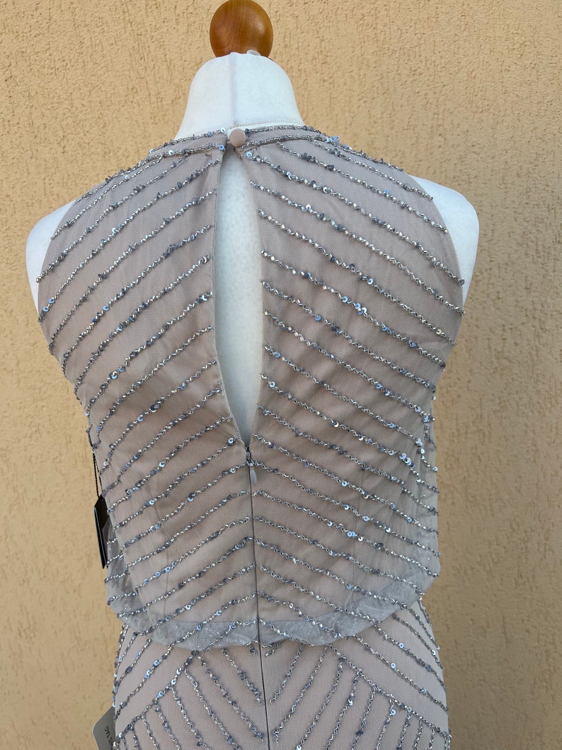 May include: A sleeveless, sequined dress in a light gray color with a geometric pattern. The dress has a keyhole back and a fitted silhouette.