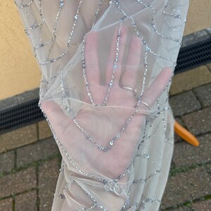 May include: Close-up of a sheer, light gray fabric with silver sequins arranged in a geometric pattern.