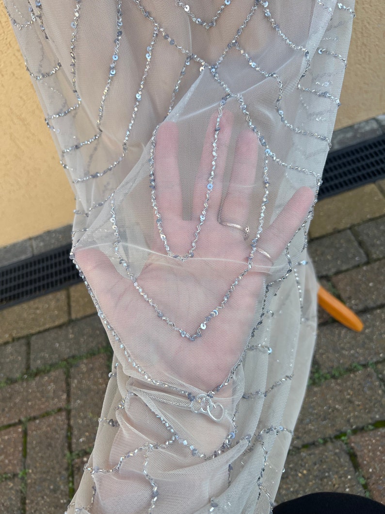 May include: Close-up of a sheer, light gray fabric with silver sequins arranged in a geometric pattern.