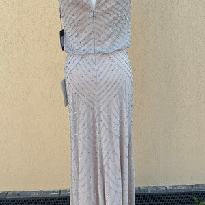 May include: A sleeveless, floor-length, beaded gown in a light gray color. The gown has a halter neckline and a keyhole back. The gown is made of a sheer fabric with a geometric pattern of beads.
