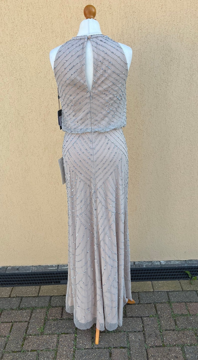 May include: A sleeveless, floor-length, beaded gown in a light gray color. The gown has a halter neckline and a keyhole back. The gown is made of a sheer fabric with a geometric pattern of beads.