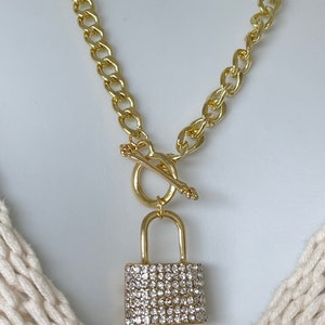 May include: Gold-tone necklace featuring a padlock pendant encrusted with clear crystals. The necklace has a chunky chain and a toggle clasp. The jewelry is displayed on a white background.