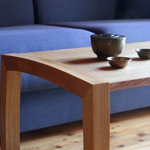 Peut inclure: Une table basse en bois avec un bord incurvé et une finition brun clair. La table a un canapé bleu en arrière-plan et trois petits bols sur le dessus.