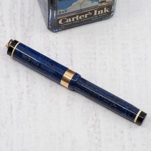May include: A blue and gold fountain pen with the text "BERTHA BARRE" on the barrel. The pen has a marbled design and gold accents. A vintage ink bottle is in the background.