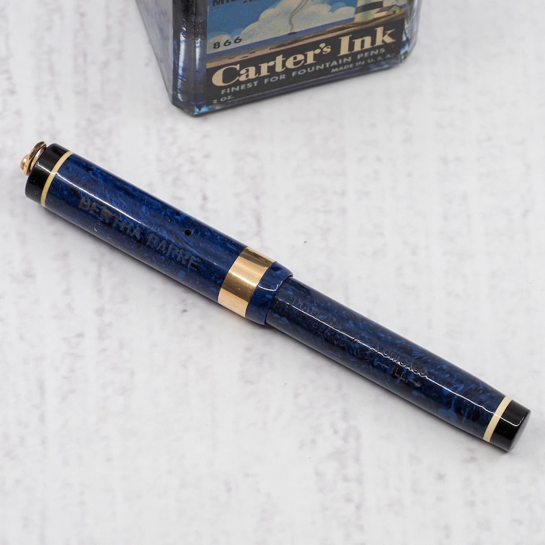 May include: A blue and gold fountain pen with the text "BERTHA BARRE" on the barrel. The pen has a marbled design and gold accents. A vintage ink bottle is in the background.