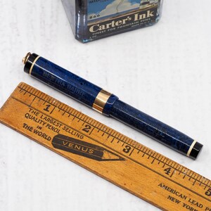 May include: A blue and gold fountain pen with the text "BERTHA BARKE" is positioned next to a wooden ruler. The ruler displays the text "THE LARGEST SELLING QUALITY PENCIL IN THE WORLD" and the brand "VENUS". A vintage ink bottle is in the background.