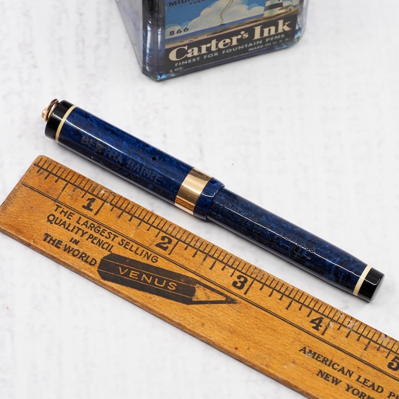 May include: A blue and gold fountain pen with the text "BERTHA BARKE" is positioned next to a wooden ruler. The ruler displays the text "THE LARGEST SELLING QUALITY PENCIL IN THE WORLD" and the brand "VENUS". A vintage ink bottle is in the background.