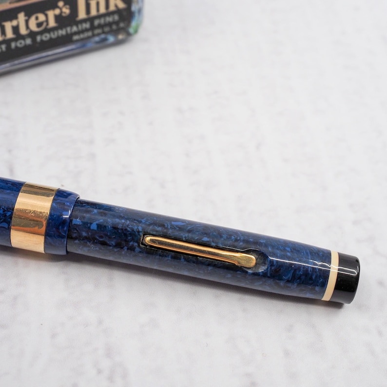 May include: Close-up of a blue and gold fountain pen. The pen has a marbled blue pattern with gold accents and a gold clip. The pen's nib is not visible. A vintage ink bottle is in the background.
