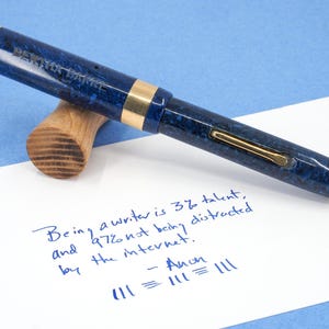 May include: A blue and gold fountain pen with a marbled design rests on a wooden pen rest. The pen is lying on a white piece of paper with handwritten text in blue ink. The text reads: "Being a writer is 3% talent, and 97% not being distracted by the internet."