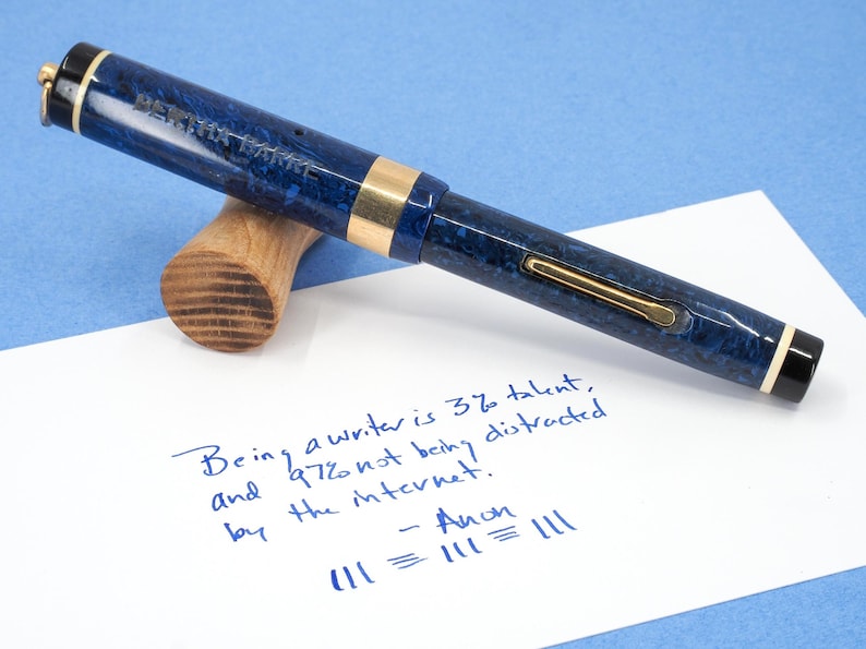 May include: A blue and gold fountain pen with a marbled design rests on a wooden pen rest. The pen is lying on a white piece of paper with handwritten text in blue ink. The text reads: "Being a writer is 3% talent, and 97% not being distracted by the internet."