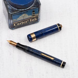 May include: A vintage fountain pen with a gold nib and a dark blue marbled barrel, alongside a bottle of "Midnight Blue" ink. The ink bottle features a lighthouse illustration and the text "Carter's Ink, Finest for Fountain Pens."