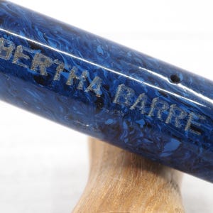 May include: Close-up of a pen with a blue and black marbled design. The pen has the name "BERTHA BARRE" engraved on the side. The pen is resting on a wooden surface.