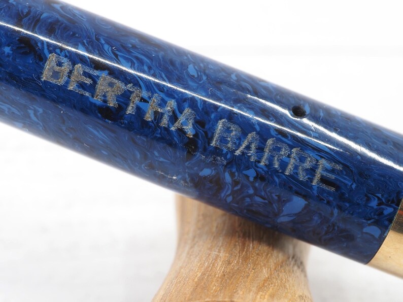 May include: Close-up of a pen with a blue and black marbled design. The pen has the name "BERTHA BARRE" engraved on the side. The pen is resting on a wooden surface.