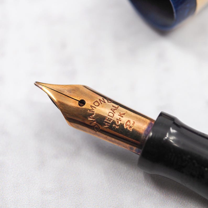 May include: Close-up of a gold-colored fountain pen nib with the text "DIAMOND MEDAL 14K". The pen has a black barrel and a blue and gold cap is visible in the background. The nib is angled towards the viewer.
