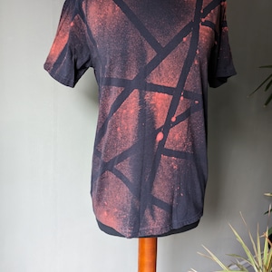 May include: Black t-shirt with a red and black geometric pattern. The pattern is created using a spray paint technique. The shirt is on a mannequin.