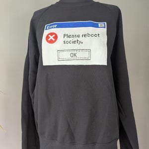 Puede incluir: Sudadera negra con un diseño gráfico blanco que simula un mensaje de error. El mensaje dice "Error" en la parte superior, con una "X" roja y el texto "Please reboot society." debajo. Un botón "OK" en la parte inferior.