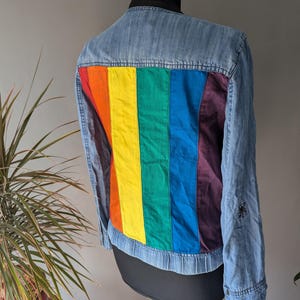 Peut inclure: Une veste en jean bleu clair avec un panneau arc-en-ciel au dos. Le panneau arc-en-ciel est composé de différentes bandes de tissu de couleurs cousues ensemble.