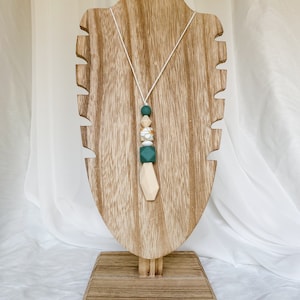 May include: A necklace with a white cord and a pendant made of various beads. The beads include green, white, and beige shapes, with a floral patterned bead. The necklace is displayed on a wooden stand.