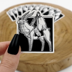 May include: Black and white sticker featuring a hooded figure riding a mythical creature with antlers. The sticker is on a wooden surface.