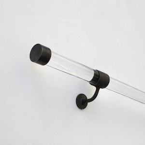 May include: A clear acrylic handrail with black metal end caps and a black metal bracket. The handrail is mounted to a white wall.