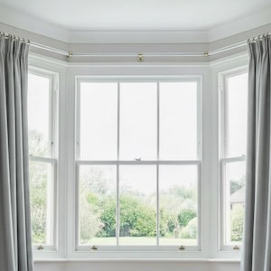 May include: A light grey window treatment with matching curtains and a gold-coloured curtain rail. The window is a bay window style with multiple panes, offering a view of lush greenery outside. The curtains are neatly pleated.
