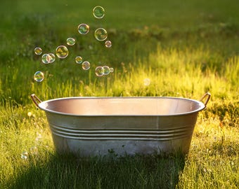 Puede incluir: Una bañera de metal plateada se encuentra en un césped verde con burbujas de jabón flotando en el aire por encima.